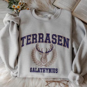 May include: A light gray sweatshirt with the word "TERRASEN" in navy blue block letters above a deer head emblem. Below the emblem is the word "GALATHYNIUS" in navy blue. The sweatshirt is folded on a white surface.