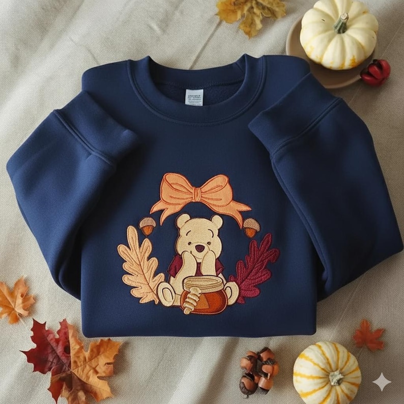 Puede incluir: Sudadera azul marino con un dise&ntilde;o bordado de un oso sosteniendo un tarro de miel, rodeado por una corona de hojas de oto&ntilde;o y bellotas. Un lazo color melocot&oacute;n est&aacute; en la parte superior. Una sudadera con tem&aacute;tica oto&ntilde;al.