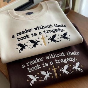 Może przedstawiać: Dwie bluzy, kremowa i brązowa, są ułożone jedna na drugiej. Każda ma wyhaftowane zdanie "a reader without their book is a tragedy". Poniżej tekstu znajdują się haftowane smoki i miecz.
