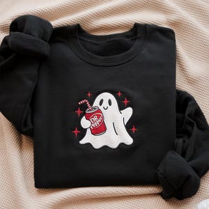 May include: Black sweatshirt with a white ghost holding a Dr. Pepper can. The ghost has a smiling face and is surrounded by four red starbursts. The Dr. Pepper can is red and white with a straw.