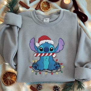 May include: A light gray sweatshirt featuring a cartoon character wearing a Santa hat and holding a candy cane. The character is surrounded by a string of colorful Christmas lights. The sweatshirt is a festive holiday item.