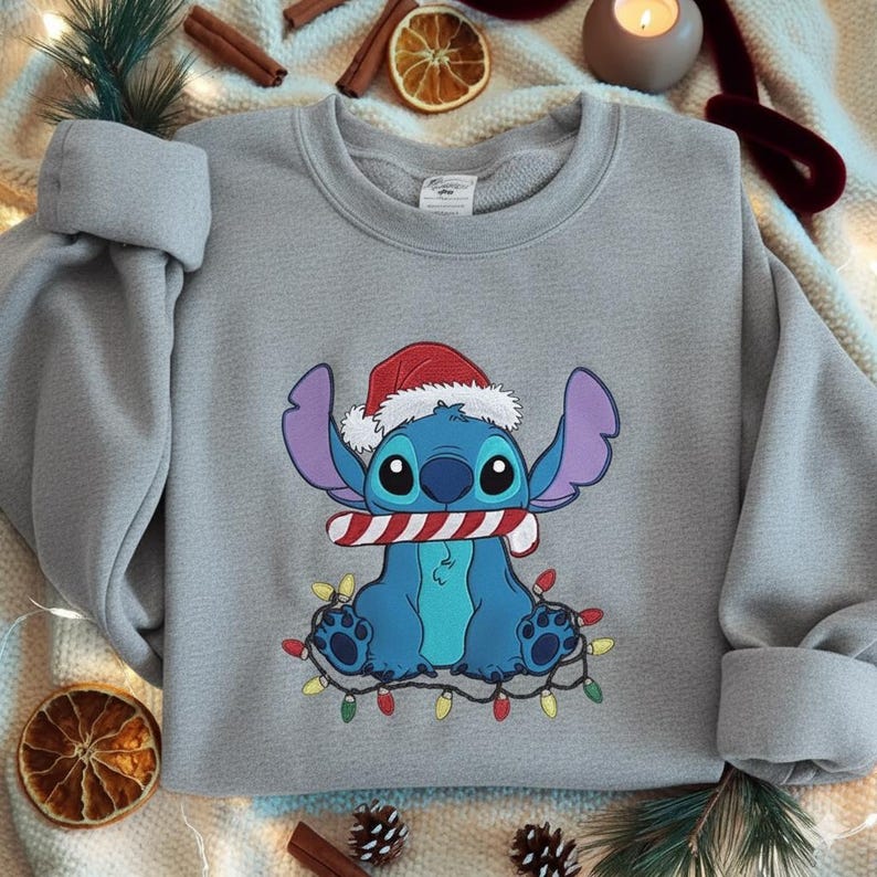 May include: A light gray sweatshirt featuring a cartoon character wearing a Santa hat and holding a candy cane. The character is surrounded by a string of colorful Christmas lights. The sweatshirt is a festive holiday item.
