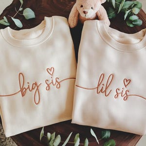 Puede incluir: Dos sudaderas color crema con las frases "big sis" y "lil sis" bordadas en escritura color cobre. Cada sudadera tiene un pequeño símbolo de corazón. Un osito de peluche y vegetación están en el fondo.