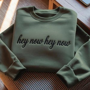 May include: An olive green crewneck sweatshirt featuring the embroidered black text "hey now hey now" across the chest. The garment has long sleeves with ribbed cuffs and a ribbed neckline, showcasing a clean, solid-colored fabric design.