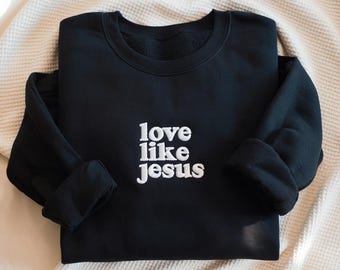 以下が含まれることがあります： 白い「love like jesus」の文字が刺繍された黒いスウェットシャツ。スウェットシャツは丸首で、袖口が折り返された長袖です。柔らかい素材で作られています。クリーム色の毛布の上に置かれています。