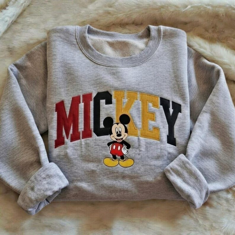 Puede incluir: Una sudadera gris con la palabra "MICKEY" en letras rojas, negras y amarillas. Una imagen de dibujos animados de Mickey Mouse est&aacute; debajo de las letras. La sudadera est&aacute; doblada y tiene pu&ntilde;os.