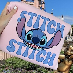 Puede incluir: Una sudadera rosa claro con el personaje Stitch de Lilo & Stitch. La palabra "STITCH" est&aacute; bordada en azul encima y debajo de la cara del personaje. El fondo incluye un castillo y una estatua.
