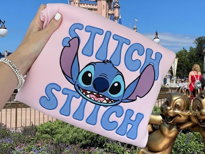 Puede incluir: Una sudadera rosa claro con el personaje Stitch de Lilo & Stitch. La palabra "STITCH" est&aacute; bordada en azul encima y debajo de la cara del personaje. El fondo incluye un castillo y una estatua.
