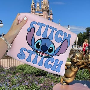 Puede incluir: Una sudadera rosa claro con un dise&ntilde;o azul de Stitch y la palabra "Stitch" en letras azules. La sudadera se sostiene frente a un castillo y una estatua de Chip y Chop.