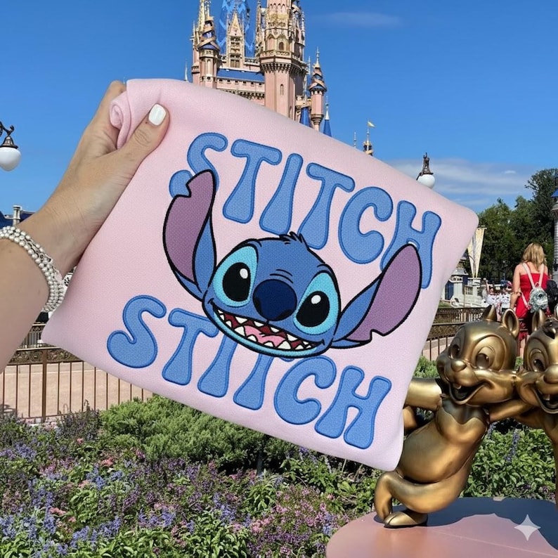 Puede incluir: Una sudadera rosa claro con un dise&ntilde;o azul de Stitch y la palabra "Stitch" en letras azules. La sudadera se sostiene frente a un castillo y una estatua de Chip y Chop.