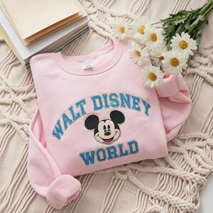 Puede incluir: Una sudadera de cuello redondo en color rosa claro con un gr&aacute;fico bordado de Mickey Mouse centrado entre el texto azul Walt Disney World. La sudadera tiene mangas largas y un cuello acanalado, con un dise&ntilde;o cl&aacute;sico en tono pastel.