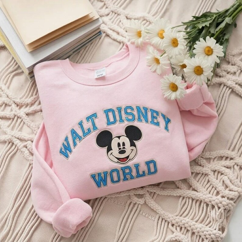 Puede incluir: Una sudadera de cuello redondo en color rosa claro con un gr&aacute;fico bordado de Mickey Mouse centrado entre el texto azul Walt Disney World. La sudadera tiene mangas largas y un cuello acanalado, con un dise&ntilde;o cl&aacute;sico en tono pastel.