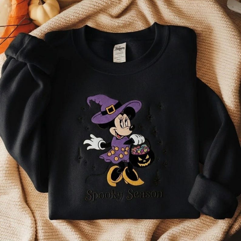 Puede incluir: Una sudadera negra de manga larga con un bordado de un rat&oacute;n de dibujos animados con un sombrero de bruja morado y un vestido con lunares amarillos. El rat&oacute;n sostiene un cubo de calabaza lleno de dulces. El texto Spooky Season est&aacute; debajo.