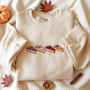 Puede incluir: Sudadera color crema con rebanadas de pastel bordadas en varios colores. Las rebanadas de pastel son marrones, amarillas y rojas, con un contorno negro. La sudadera tiene mangas largas y cuello redondo. Tema otoñal.