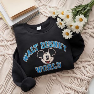 Puede incluir: Una sudadera negra de cuello redondo con un gr&aacute;fico de Mickey Mouse centrado entre las palabras WALT DISNEY en una fuente azul arqueada y la palabra WORLD en una fuente azul recta, con contornos blancos alrededor de las letras.