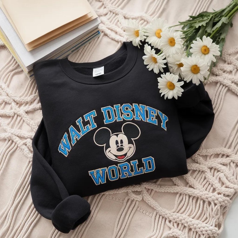 Puede incluir: Una sudadera negra de cuello redondo con un gr&aacute;fico de Mickey Mouse centrado entre las palabras WALT DISNEY en una fuente azul arqueada y la palabra WORLD en una fuente azul recta, con contornos blancos alrededor de las letras.