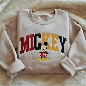 Puede incluir: Sudadera beige con la palabra "MICKEY" en letras rojas, negras y amarillas. Una imagen de dibujos animados de Mickey Mouse est&aacute; centrada debajo de las letras. La sudadera tiene cuello redondo y mangas largas con pu&ntilde;os doblados.