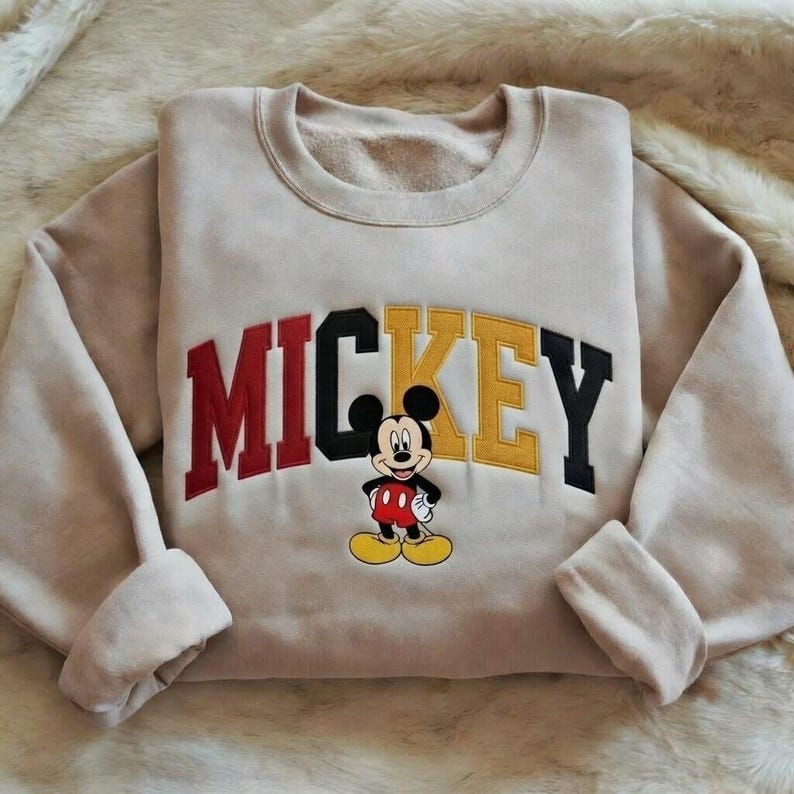 Puede incluir: Sudadera beige con la palabra "MICKEY" en letras rojas, negras y amarillas. Una imagen de dibujos animados de Mickey Mouse est&aacute; centrada debajo de las letras. La sudadera tiene cuello redondo y mangas largas con pu&ntilde;os doblados.