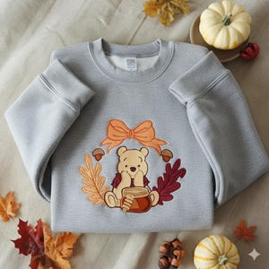 Puede incluir: Una sudadera gris claro con un dise&ntilde;o bordado de un oso con un tarro de miel, rodeado por una corona de hojas de oto&ntilde;o, bellotas y un lazo naranja. La sudadera se muestra sobre una superficie neutra con decoraciones oto&ntilde;ales.