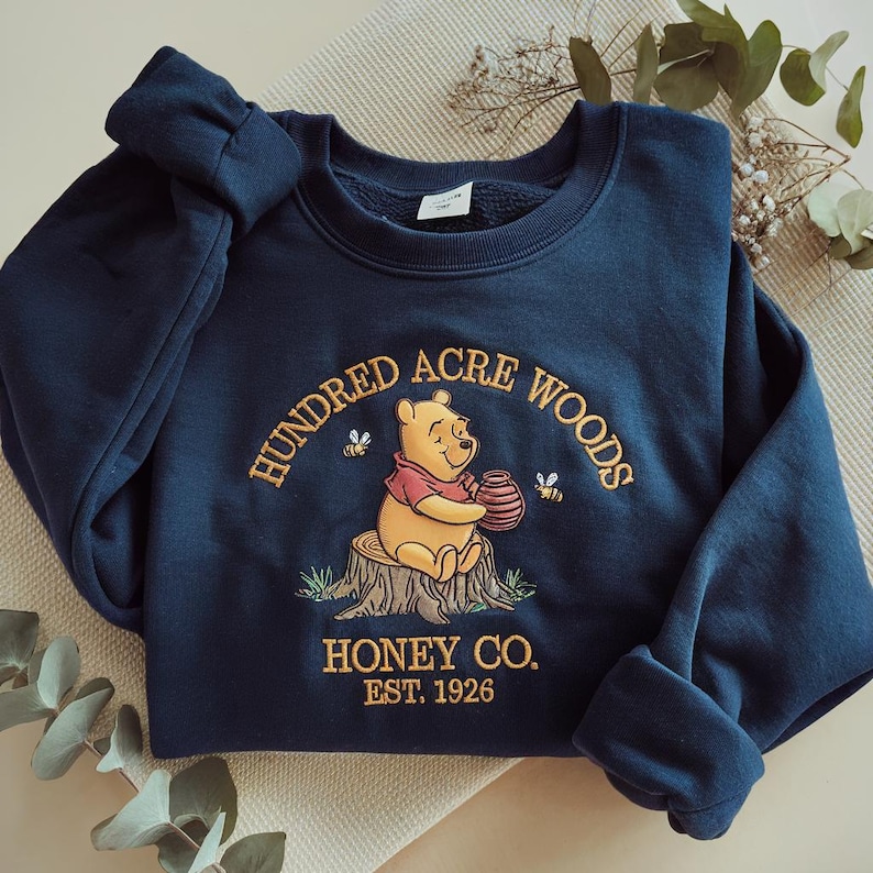 Puede incluir: Sudadera azul marino con un dise&ntilde;o bordado. Presenta a Winnie the Pooh sosteniendo un tarro de miel, con el texto "Hundred Acre Woods Honey Co. Est. 1926". El dise&ntilde;o incluye abejas y un toc&oacute;n de &aacute;rbol.