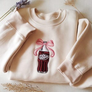 Peut inclure: Un sweat-shirt beige avec un col rond et des manches longues. Il présente une bouteille de Dr. Pepper brodée avec un nœud rose. La bouteille de Dr. Pepper est rouge, blanche et noire. Le sweat-shirt est plié sur une surface blanche.