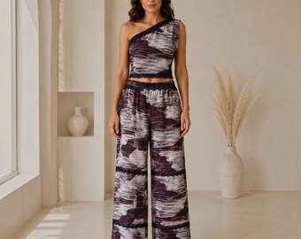 May include: A two-piece set consisting of a one-shoulder crop top and wide-leg trousers. The garments feature a dark purple, grey, and white abstract print with black lace trim detailing along the neckline, hem, and horizontal leg panels.