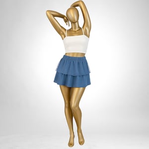 May include: A white crop top with spaghetti straps and a blue tiered mini skirt. The skirt has a dotted pattern and a sheer layer at the hem. The mannequin is gold-colored and posed with arms overhead.