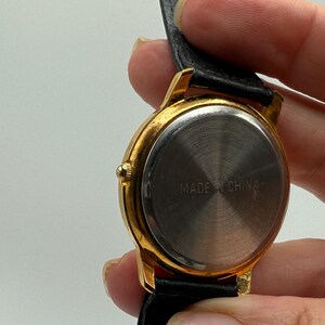May include: Close-up of a gold-toned wristwatch with a black leather band. The watch face is silver, and the back of the watch has the text "MADE IN CHINA". The watch is held by a person.