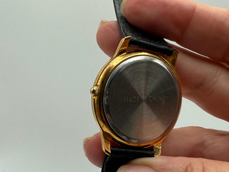 May include: Close-up of a gold-toned wristwatch with a black leather band. The watch face is silver, and the back of the watch has the text "MADE IN CHINA". The watch is held by a person.