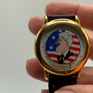 May include: Gold-tone wristwatch with a black leather band. The watch face features a cartoon illustration set against a backdrop of the American flag. The watch has a gold bezel and a black leather strap.