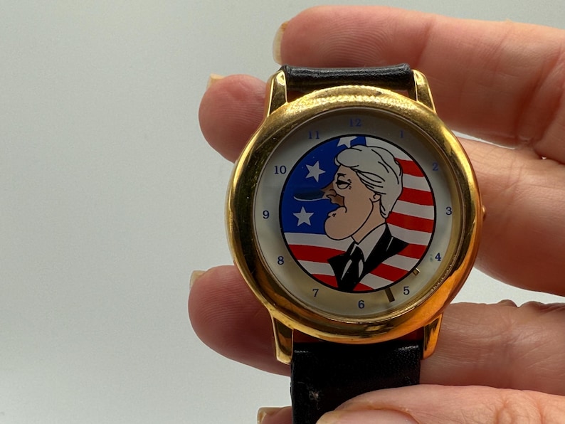 May include: Gold-tone wristwatch with a black leather band. The watch face features a cartoon illustration set against a backdrop of the American flag. The watch has a gold bezel and a black leather strap.
