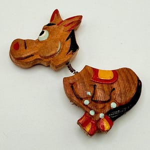 May include: A wooden bobblehead horse pin featuring a carved head and body connected by a metal spring. The horse is painted in brown with red, yellow, black, and light blue accents, including a saddle detail on the back and a red nose.