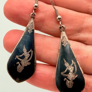 May include: A pair of teardrop-shaped dangle earrings featuring a dark blue enamel inlay with a silver-toned engraved figure of a dancer. The earrings have silver-toned metal hooks and a small decorative silver-toned emblem at the top of each piece.