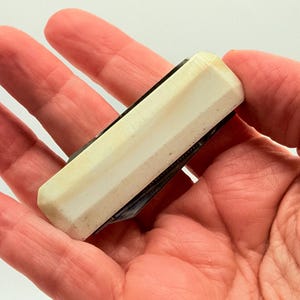 May include: A small, rectangular pocket knife with a cream-colored, ridged handle. The knife is held in an open palm, showcasing its compact size and design. The blade is not visible.