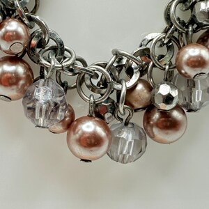 May include: Close-up of a silver-toned charm bracelet with various dangling charms. The charms include faux pearls in a soft pink hue, faceted clear beads, and metallic beads. The bracelet is on a white background.