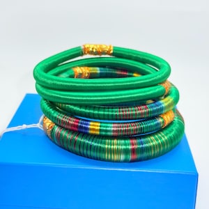 May include: A stack of green and multicolored bangles. The top bangles are solid green, while the lower ones feature stripes of red, blue, and yellow. The bangles are resting on a blue box, creating a vibrant contrast. The bangles have gold accents.