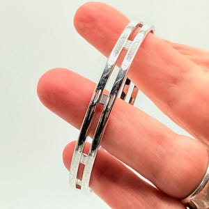 May include: A silver-colored bangle bracelet with a rectangular cut-out design. The bracelet is held between fingers, showcasing its polished, reflective surface. The jewelry piece is a simple, elegant accessory.