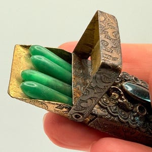 May include: An antique silver match safe with a hinged lid, showcasing intricate embossed patterns. The open compartment reveals four vibrant green matchsticks. A small, oval-shaped blue stone adorns the side, adding a touch of elegance.