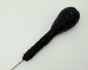 May include: A black beaded pin with a rounded top and a long, textured handle. The pin is made of small, dark beads, and the handle tapers to a fine, silver-colored point. The pin is set against a white background.