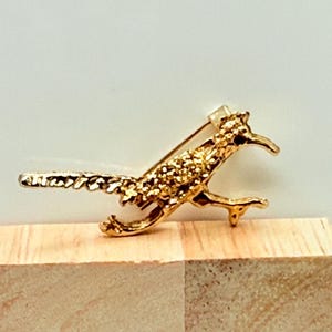 May include: A gold-colored roadrunner bird pin. The pin is detailed with textured feathers and a curved beak. The bird is depicted in a running pose, with its legs extended. The pin has a clasp on the back for secure attachment.