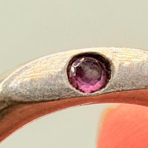 May include: A silver-toned metal ring featuring a single, small, round, faceted magenta-colored gemstone set flush into the center of the band. The band has a smooth, polished finish with a slightly tapered shape around the stone setting.