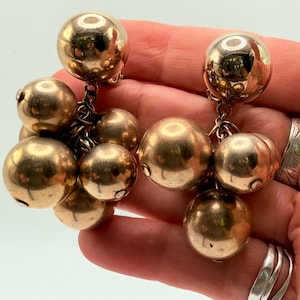 May include: A pair of gold-toned ball drop earrings. Each earring features a cluster of various-sized spheres suspended from a chain. The earrings have a polished finish, reflecting light. The jewelry is held in a hand.