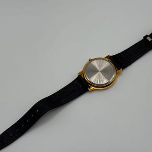 May include: A gold-toned wristwatch with a black leather band. The watch face is round, and the back of the watch is visible, showing a silver-colored metal. The band has a gold-colored buckle.