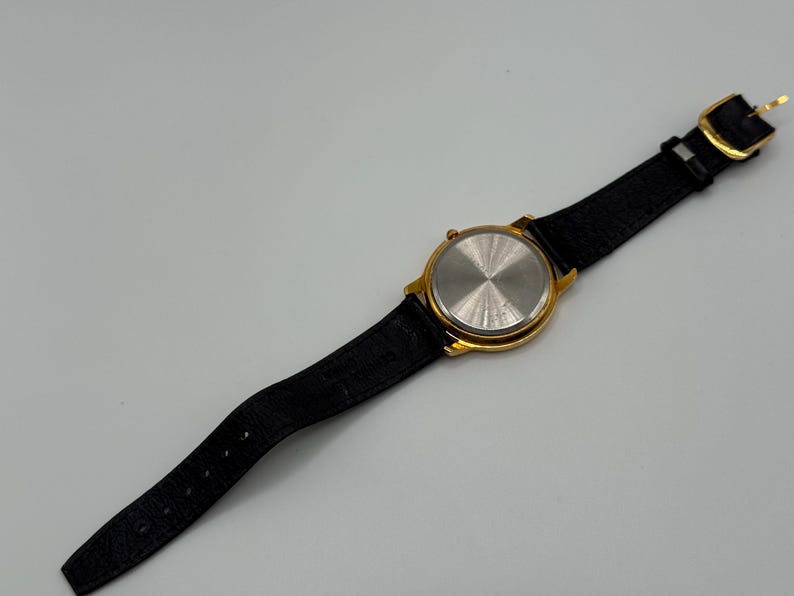 May include: A gold-toned wristwatch with a black leather band. The watch face is round, and the back of the watch is visible, showing a silver-colored metal. The band has a gold-colored buckle.