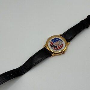 May include: Gold-tone wristwatch with a black leather band. The watch face features a patriotic design with red and white stripes, blue stars, and a cartoon character. The watch is a classic accessory.