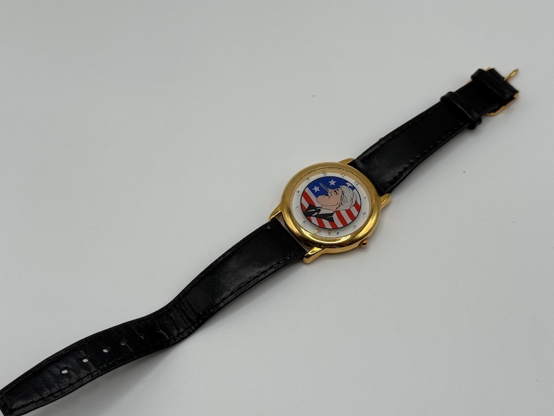 May include: Gold-tone wristwatch with a black leather band. The watch face features a patriotic design with red and white stripes, blue stars, and a cartoon character. The watch is a classic accessory.