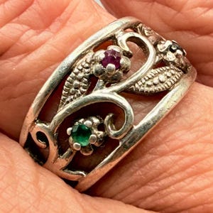 May include: A silver ring featuring an openwork design with swirling metal patterns, textured leaf motifs, and three small inset gemstones: one round red stone, one round green stone, and one round dark blue stone set within the metal frame.