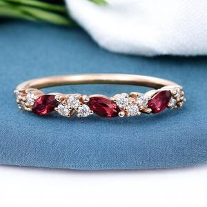 May include: A rose gold ring with alternating red and white gemstones. The ring features oval-shaped red stones and smaller, round white stones. The text "SANIYABYJEWELRY" is visible in the upper right corner.