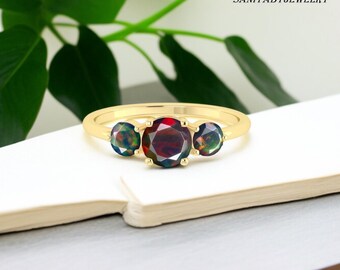 May include: A gold ring featuring three gemstones. The center stone is a deep red, flanked by two smaller, iridescent stones. The ring is set on a white surface with green foliage in the background. The text "SANIYABYJEWELRY" is visible.