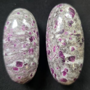 Può includere: Due pietre levigate di forma ovale caratterizzate da una matrice grigia e bianca maculata, punteggiata da inclusioni di rubino color magenta vibrante. Le pietre presentano una finitura liscia e lucida che mette in risalto il contrasto tra la base minerale scura e i cristalli rosa brillante incastonati sulla superficie di ogni pezzo.